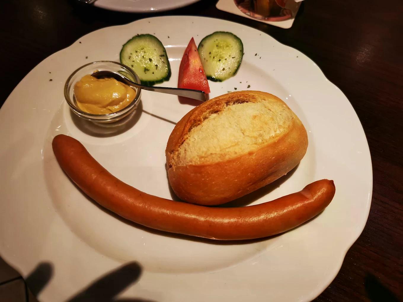 Ein Brötchen und eine Wurst auf einem Teller, garniert mit Gemüse und Senf