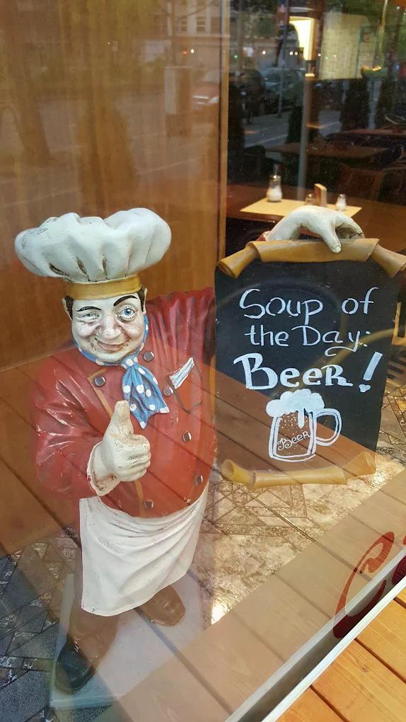 Eine fröhliche Kochfigur mit rotem Mantel und weißer Kochmütze hält ein Bier-Schild