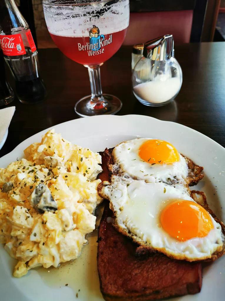 Zwei Spiegeleier mit Bratfleisch und Kartoffelsalat auf weißem Teller, dazu Getränk