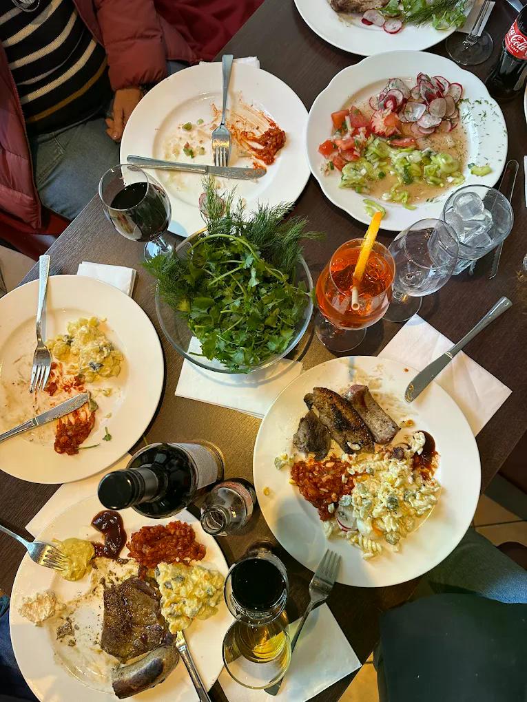 Fünf Teller mit gegrilltem Fleisch, Kartoffelsalat und Gemüse, dazu Getränke und Besteck auf einem dunklen Holztisch