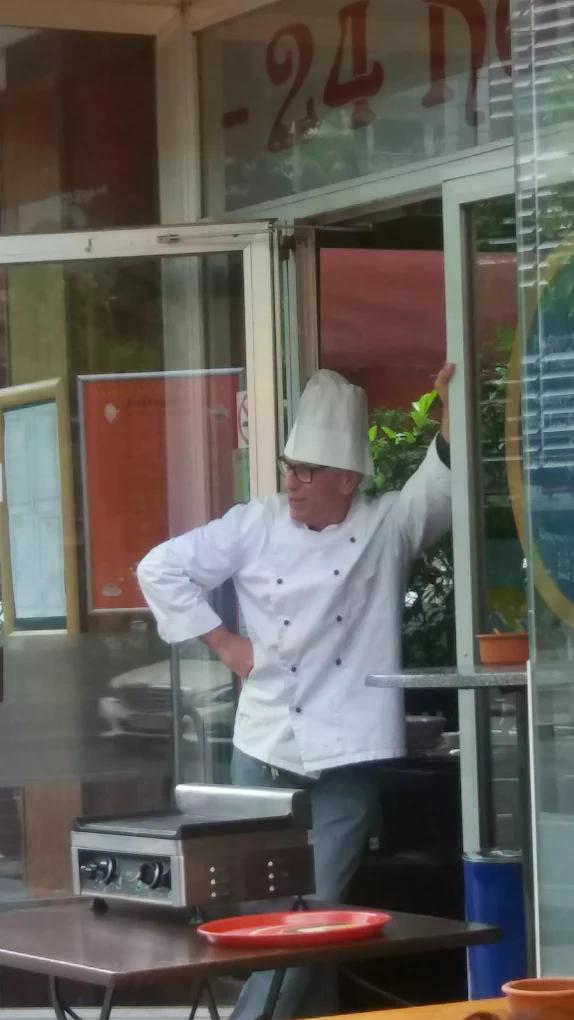 Ein älterer Koch mit weißer Kochmütze lächelt entspannt im Eingang eines Restaurants